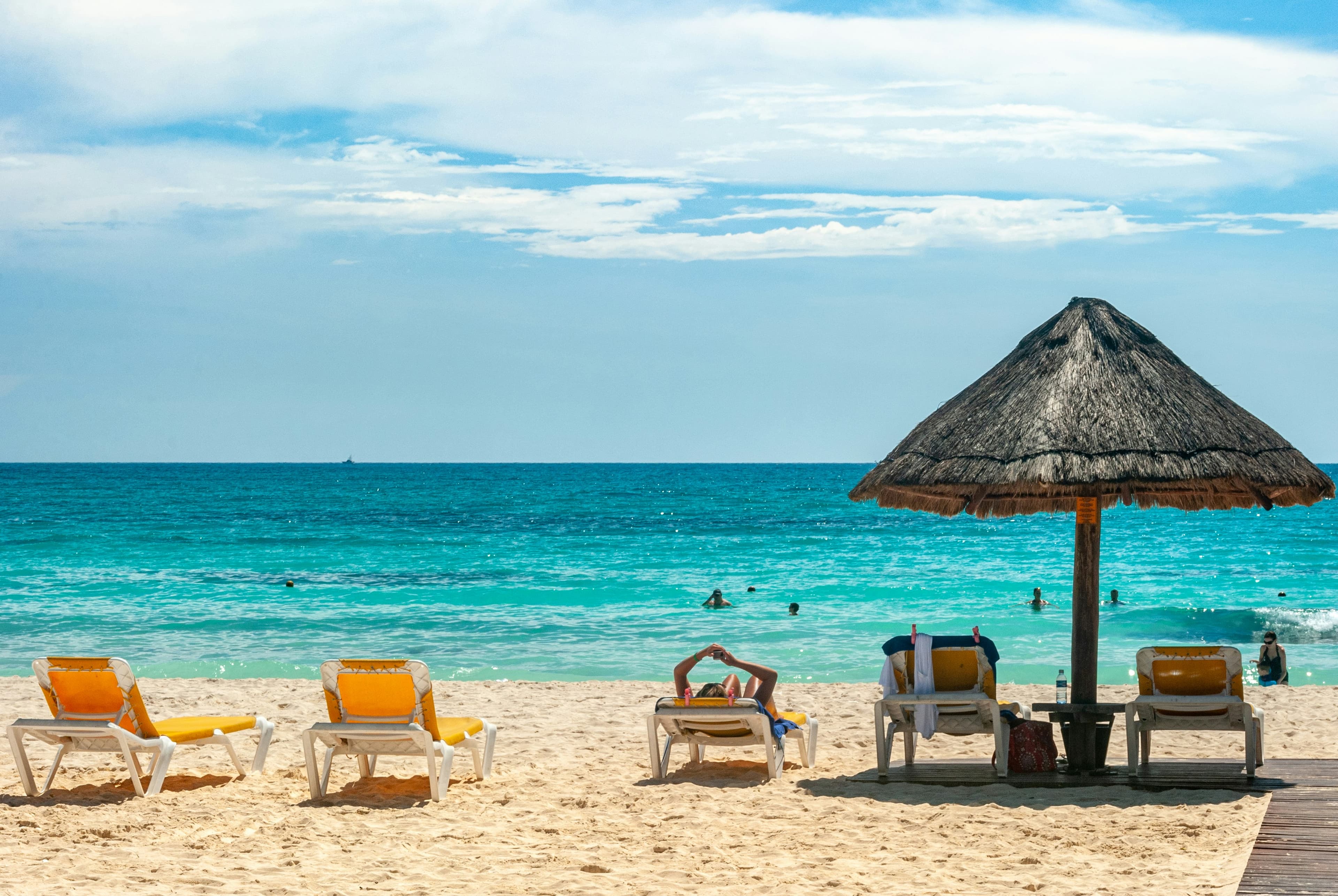 Yellow sun loungers and a thatched parasol on a sandy beach overlooking turquoise water, with swimmers in the sea beneath a bright, cloudy sky.