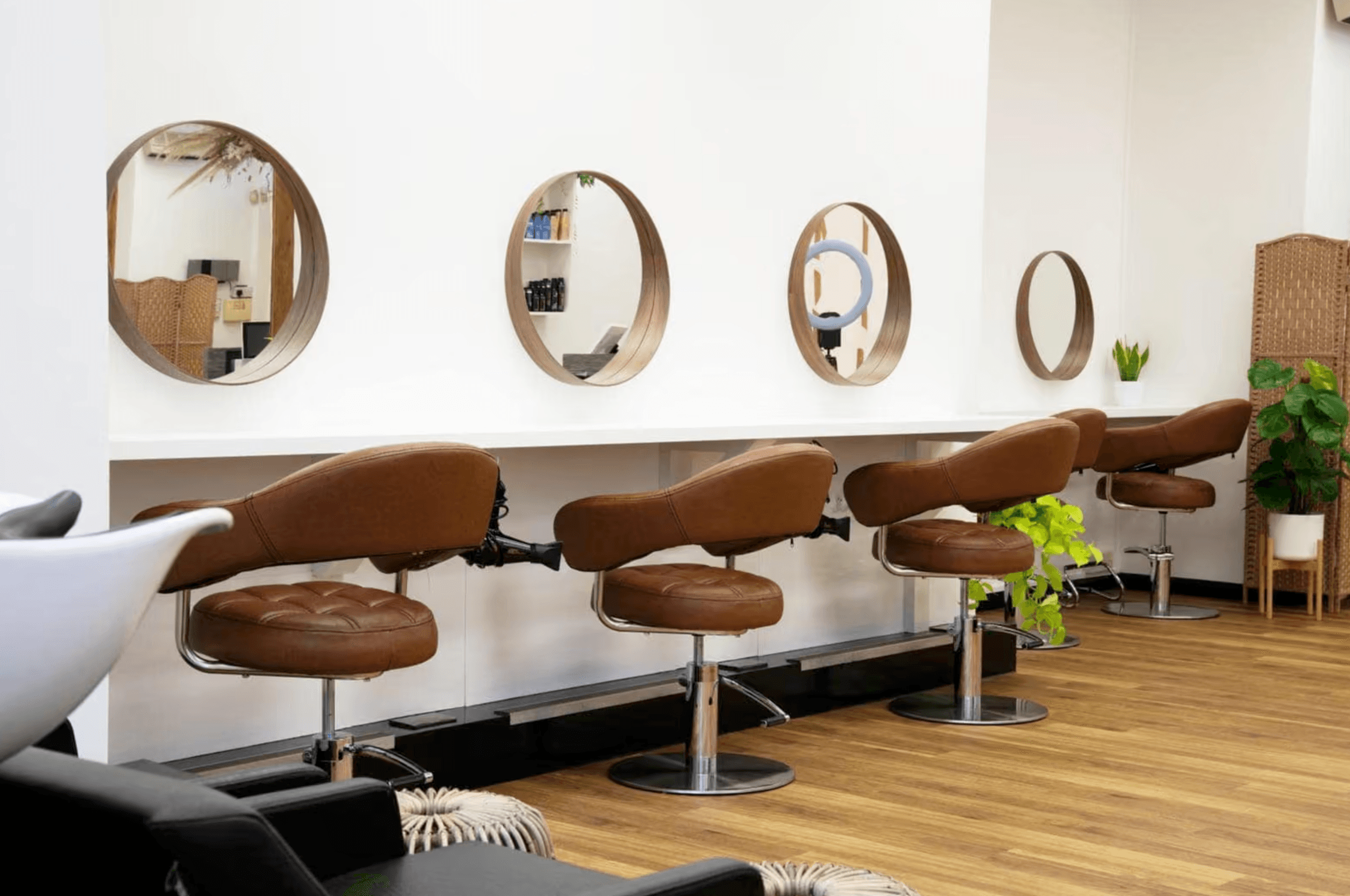 A row of brown leather salon chairs sits beneath round wooden-framed mirrors along a white wall, with a wash basin and potted plants nearby.